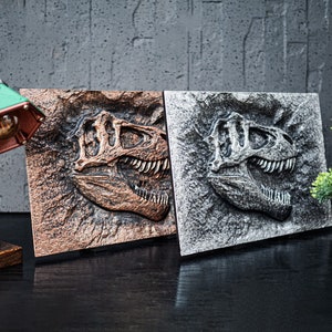 Può includere: Due pezzi di arredamento da parete a forma di cranio di dinosauro, uno in finitura rame e l'altro in finitura argento. I crani sono montati su uno sfondo strutturato.