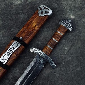 May include: A sword with a patterned Damascus steel blade, a wooden handle with silver-colored metal accents, and a matching wooden scabbard featuring silver-colored metal fittings with intricate knotwork designs.
