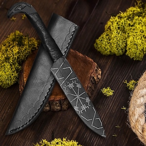 May include: A black knife with a decorative handle and a sheath. The knife blade features a silver geometric design. The handle is shaped like a bird's head. The knife and sheath are on a wooden surface with moss.