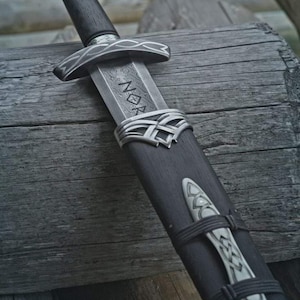 May include: A sword with a dark wooden handle and a silver pommel featuring intricate engravings. The blade displays a visible pattern-welded texture and the letters NOR etched into the metal. It is housed in a dark wooden scabbard with silver metal accents and decorative knotwork patterns.