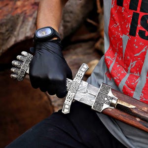 Op de afbeelding: Een zwaard met een patroon van Damascusstaal, een zilverkleurig metalen gevest met ingewikkelde Keltische knoopmotieven en een vuistvormige pommel. Het zwaard zit gedeeltelijk in een donkerbruine houten schede.