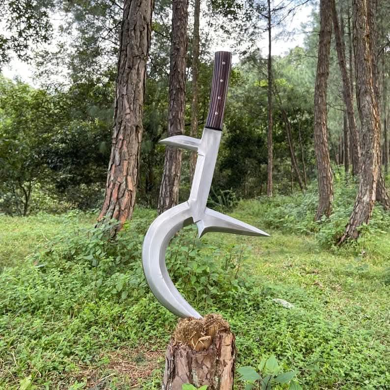 May include: A large, silver-colored, curved blade with a wooden handle. The blade has a unique shape with multiple points and is displayed outdoors, resting on a tree stump. The handle has a dark brown and light brown striped pattern.