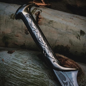 May include: A metallic sword with a dark, polished finish featuring intricate, engraved vine-like patterns along the handle and the base of the blade. The design includes a curved pommel and a sharp, tapered edge, showcasing detailed metalwork.