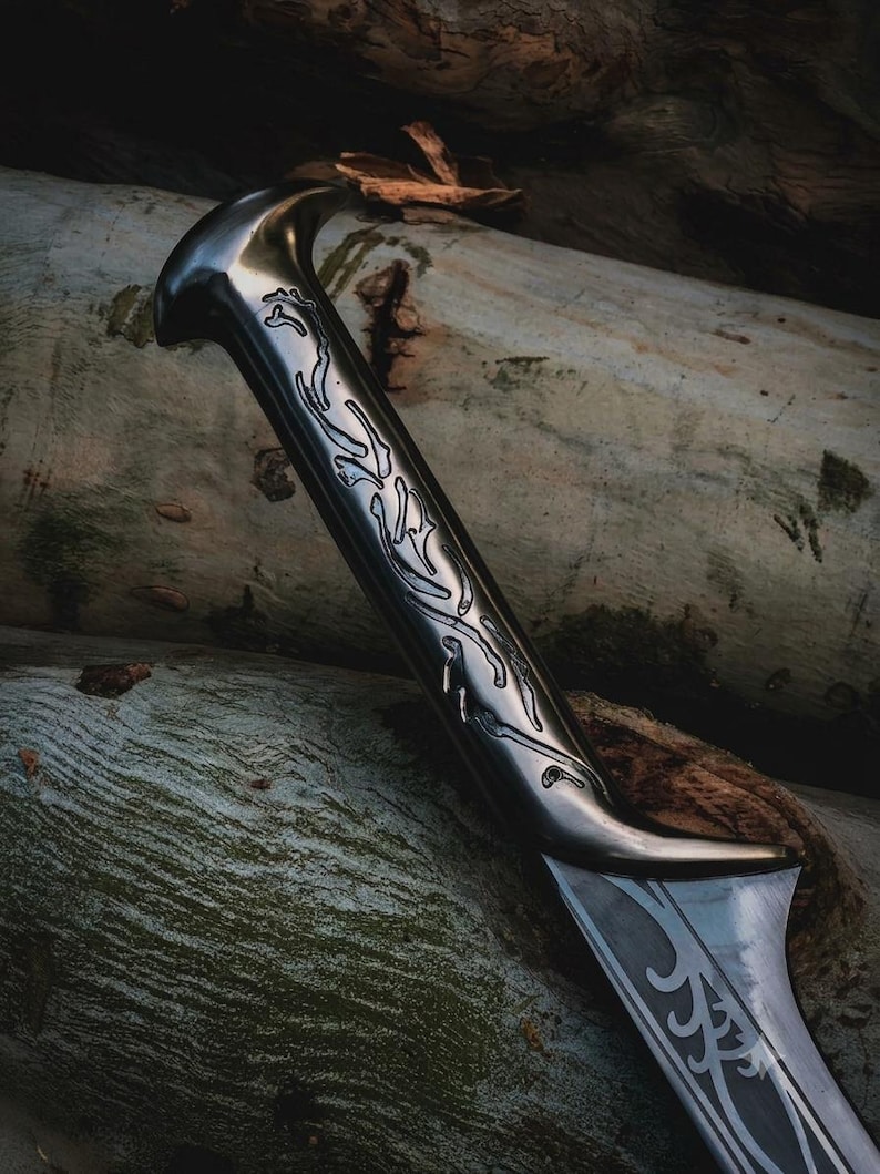 May include: A metallic sword with a dark, polished finish featuring intricate, engraved vine-like patterns along the handle and the base of the blade. The design includes a curved pommel and a sharp, tapered edge, showcasing detailed metalwork.