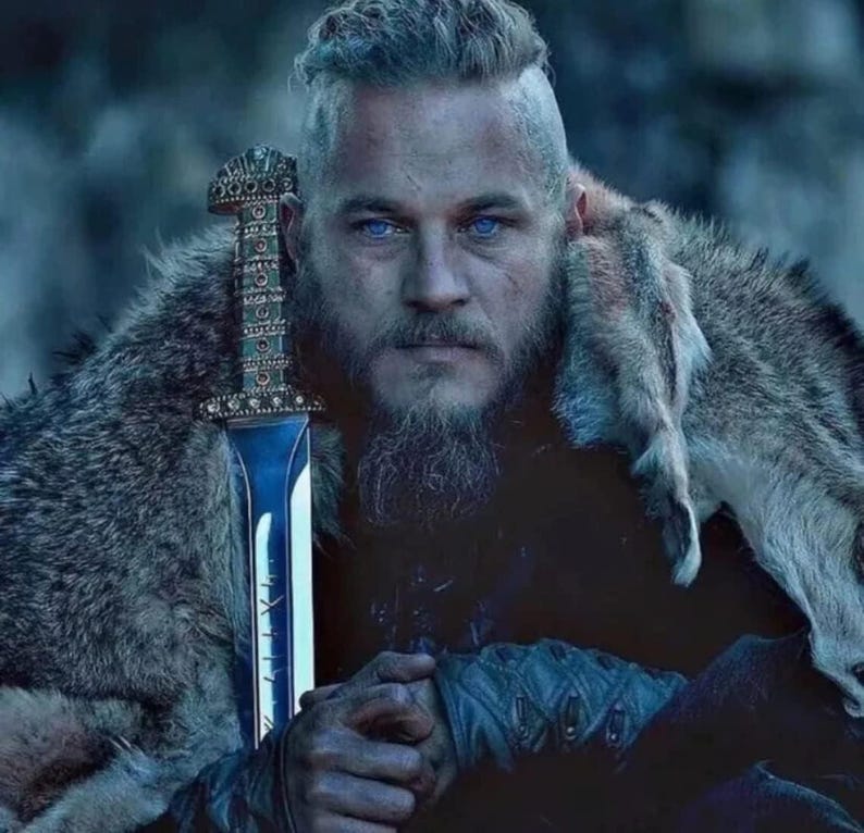 May include: A person with a shaved side hairstyle and a full beard, wearing a textured fur garment over the shoulders. A metallic sword with an intricate, patterned hilt and a polished blade is held upright in front of the chest.