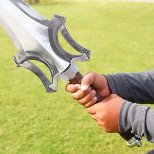 Op de afbeelding: Een zilverkleurig metalen zwaard met een breed, tweesnijdend lemmet en een complexe, opengewerkte stootplaat. Het handvat is omwikkeld met bruin materiaal en de pommel is een eenvoudige, afgeronde zilverkleurige metalen dop aan de basis.