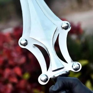Op de afbeelding: Een zilverkleurig metalen zwaardblad met een centrale richel en een decoratieve pareerstang met vier afgeronde, gepolijste metalen klinknagels. Het blad heeft een symmetrische, puntige vorm met gebogen uitsparingen aan beide zijden.