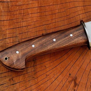 May include: Close-up of a knife with a dark brown wooden handle. The handle features three white dot inlays and a brass bolster. The blade is silver and the knife is set against a wooden background.