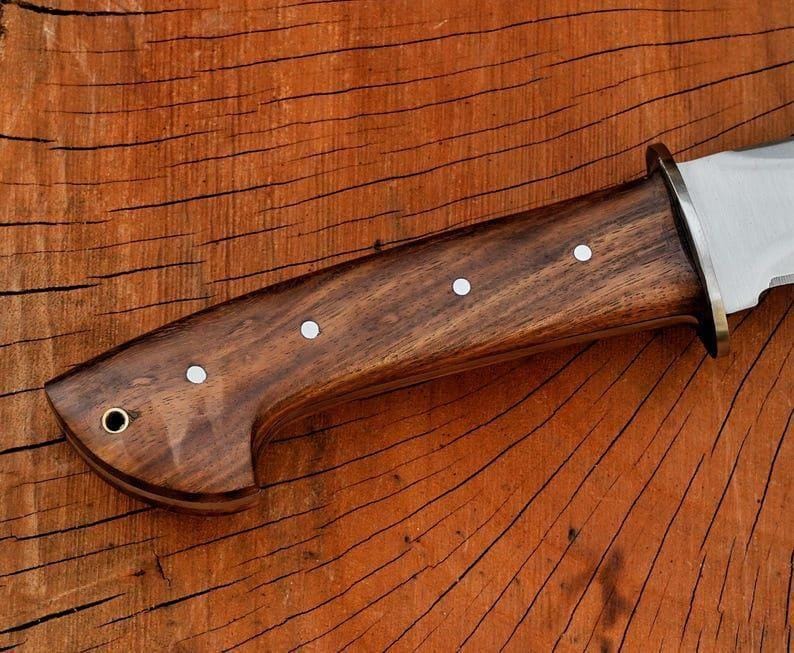 May include: Close-up of a knife with a dark brown wooden handle. The handle features three white dot inlays and a brass bolster. The blade is silver and the knife is set against a wooden background.