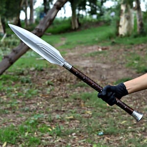 May include: A spear with a polished silver blade and a dark brown wooden handle. The handle is wrapped with a lighter brown leather and silver accents. The spear is held against a blurred green background.