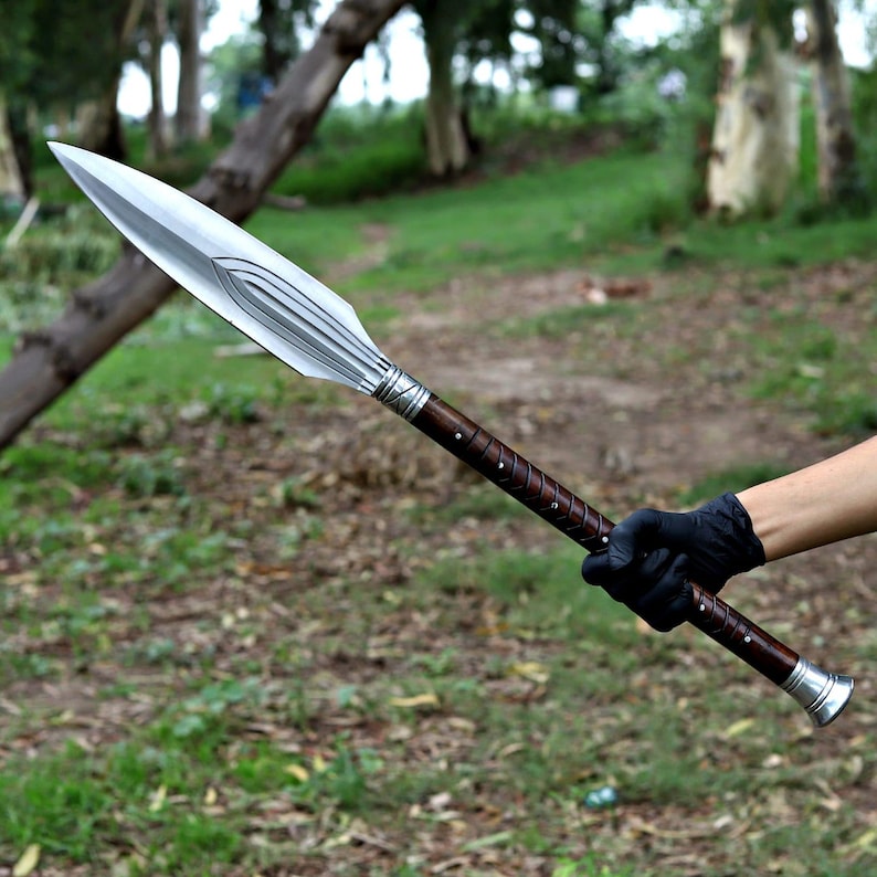 May include: A spear with a polished silver blade and a dark brown wooden handle. The handle is wrapped with a lighter brown leather and silver accents. The spear is held against a blurred green background.