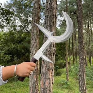 May include: A silver-colored, curved blade weapon with a wooden handle. The blade has a unique, angular design with a sharp, crescent-shaped edge. The handle is dark brown with a textured grip. The weapon is held in front of a forest background.