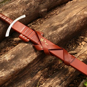 May include: A sword with a brown leather-wrapped hilt and a silver pommel featuring a circular emblem. The silver crossguard is engraved with the word CHRISTUS. The brown leather scabbard is adorned with gold-colored cross symbols and straps.