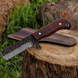 May include: A handcrafted knife with a dark wood handle and a textured, patterned blade. The knife rests on a tree stump with a brown leather sheath. The blade has a unique design.