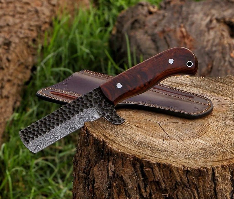 May include: A handcrafted knife with a dark wood handle and a textured, patterned blade. The knife rests on a tree stump with a brown leather sheath. The blade has a unique design.