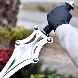 Op de afbeelding: Een zilverkleurige metalen dolk met een breed, puntig lemmet met decoratieve uitsparingen en vier opvallende ronde klinknagels. Het handvat is zwart met een metalen pommel, vastgehouden door een hand met een zwarte handschoen.
