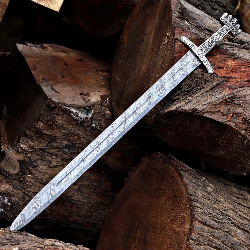 Op de afbeelding: Een tweesnijdend zwaard met een zilverkleurig lemmet met een zichtbaar golvend Damascus-staalpatroon. Het gevest bevat een pareerstang en een pommel, beide versierd met ingewikkelde, gegraveerde knoopmotieven in een metalen afwerking.