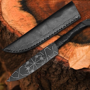 May include: A black, handcrafted knife with a leather sheath, resting on a wooden surface. The knife features a bird-shaped handle and etched geometric designs on the blade. A dried leaf is next to the knife and sheath.