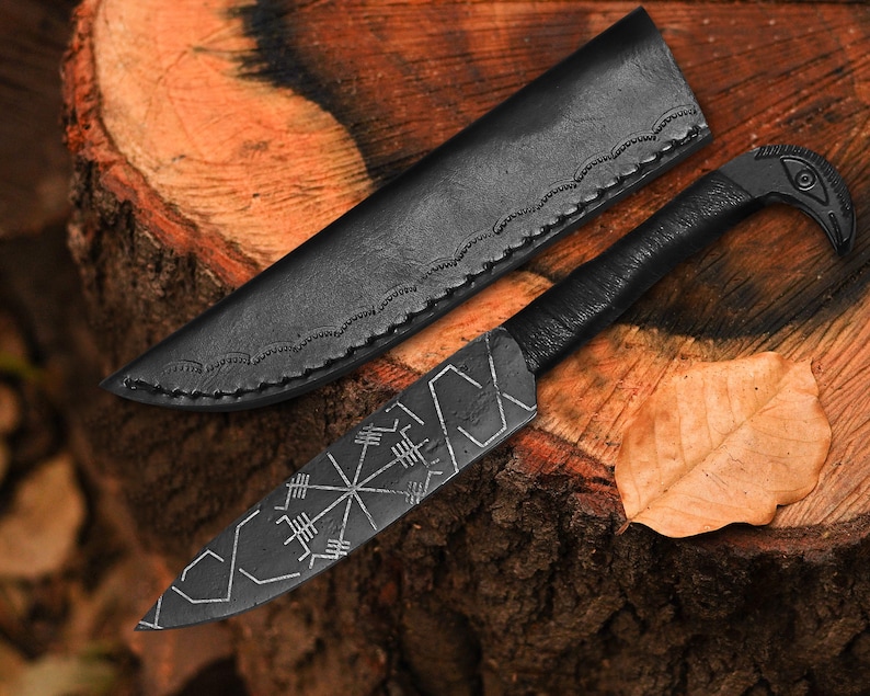 May include: A black, handcrafted knife with a leather sheath, resting on a wooden surface. The knife features a bird-shaped handle and etched geometric designs on the blade. A dried leaf is next to the knife and sheath.