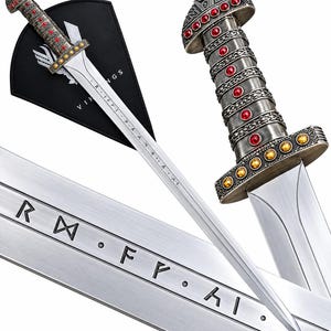 Op de afbeelding: Een zilverkleurig zwaard met een metalen gevest versierd met rode en gele ronde inlays. Het lemmet toont gegraveerde runentekens. Een zwarte driehoekige schede met de witte tekst VIKINGS is op de achtergrond zichtbaar.