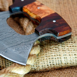 May include: A close-up of a  handmade,  damascus steel cleaver with a wooden handle. The handle is made of two different types of wood, one light and one dark. The cleaver is resting on a burlap surface.