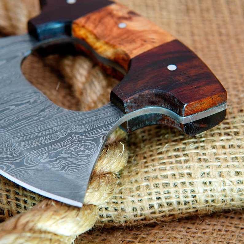 May include: A close-up of a  handmade,  damascus steel cleaver with a wooden handle. The handle is made of two different types of wood, one light and one dark. The cleaver is resting on a burlap surface.