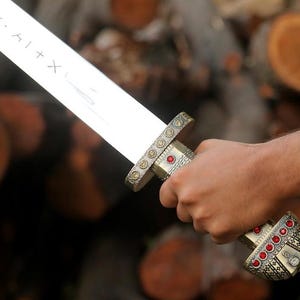May include: A silver-toned sword featuring a blade with etched runic symbols. The hilt is crafted from metallic material with intricate patterns, accented by small circular red gemstones embedded along the grip and the rounded pommel base.