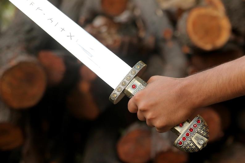 May include: A silver-toned sword featuring a blade with etched runic symbols. The hilt is crafted from metallic material with intricate patterns, accented by small circular red gemstones embedded along the grip and the rounded pommel base.