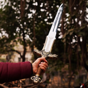 May include: A double-edged sword with a polished silver blade and a detailed bronze-colored hilt. The crossguard features ornate animal head designs, while the pommel is shaped like a skull with horns. The grip is wrapped in a textured brown material.