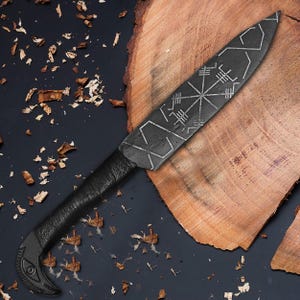 May include: A black, handcrafted knife with a textured blade featuring etched geometric designs. The handle is wrapped in black material, and the curved end has an eye-like detail. The knife rests on a wooden surface.