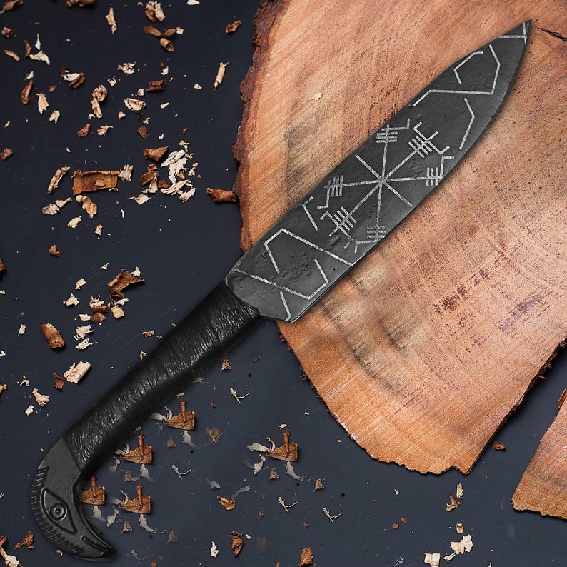 May include: A black, handcrafted knife with a textured blade featuring etched geometric designs. The handle is wrapped in black material, and the curved end has an eye-like detail. The knife rests on a wooden surface.