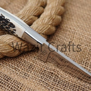 May include: A handcrafted knife with a polished steel blade and a handle featuring a cream-colored section with dark brown and black patterns. The knife is set against a backdrop of a thick, coiled rope and burlap.