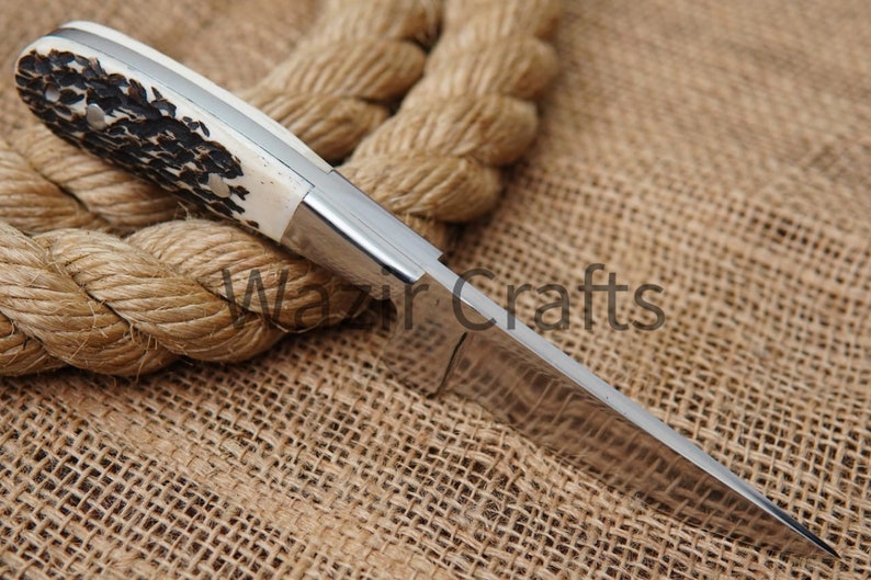 May include: A handcrafted knife with a polished steel blade and a handle featuring a cream-colored section with dark brown and black patterns. The knife is set against a backdrop of a thick, coiled rope and burlap.