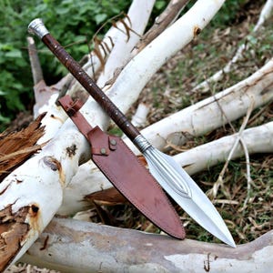 May include: A handcrafted spear with a polished silver blade and a dark brown wooden shaft wrapped in leather. The spear is accompanied by a brown leather sheath. The spear is resting on white tree branches.