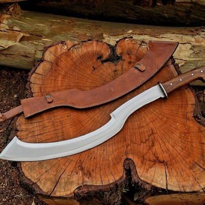 May include: A large, curved blade knife with a wooden handle and a leather sheath. The blade is silver, and the handle is a dark brown. The sheath is a lighter brown. The knife is resting on a wooden surface.