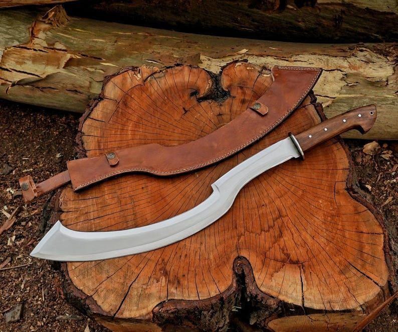 May include: A large, curved blade knife with a wooden handle and a leather sheath. The blade is silver, and the handle is a dark brown. The sheath is a lighter brown. The knife is resting on a wooden surface.