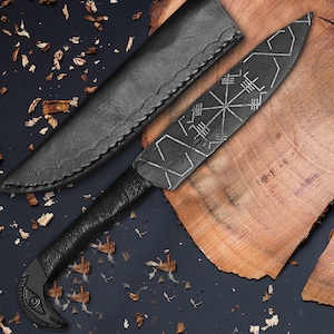 May include: A black, handcrafted knife with a leather sheath. The blade features etched geometric designs and a wrapped handle. The knife is displayed on a wooden surface, showcasing its intricate details and craftsmanship.