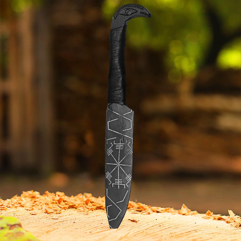 May include: A black knife with a bird-shaped handle and a blade etched with geometric designs. The handle is wrapped in black material. The knife is displayed on a wooden surface.