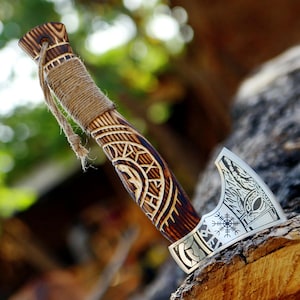 May include: A handcrafted axe with a wooden handle featuring intricate carved patterns and a wrapped grip. The silver-colored blade displays a detailed design. The axe is a decorative item.