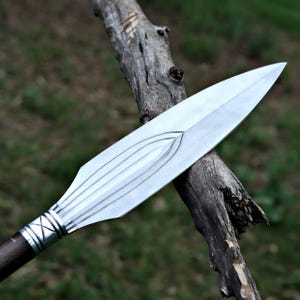 May include: A spearhead with a polished, silver blade featuring a series of parallel grooves. The spear has a dark wooden handle with silver accents. The spear is resting on a weathered, gray tree branch.