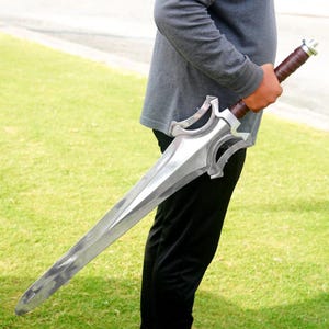 Op de afbeelding: Een groot metalen zwaard met een tweesnijdend lemmet, een complexe zilverkleurige stootplaat met gebogen uitsparingen en een bruin, met leer omwikkeld handvat met een metalen pommel, vastgehouden tegen een grijs shirt en een zwarte broek.