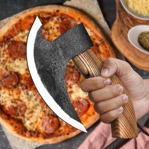 Op de afbeelding: Een zwarte en zilveren pizzames met een houten handvat. De mes is gevormd als een kleine bijl en heeft een gebogen lemmet. De mes wordt door een hand boven een pizza gehouden.