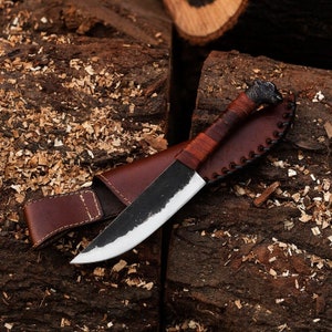 May include: A handcrafted knife with a dark blade and a light-colored edge, featuring a wrapped leather handle and a decorative animal head at the end. The knife rests in a brown leather sheath.