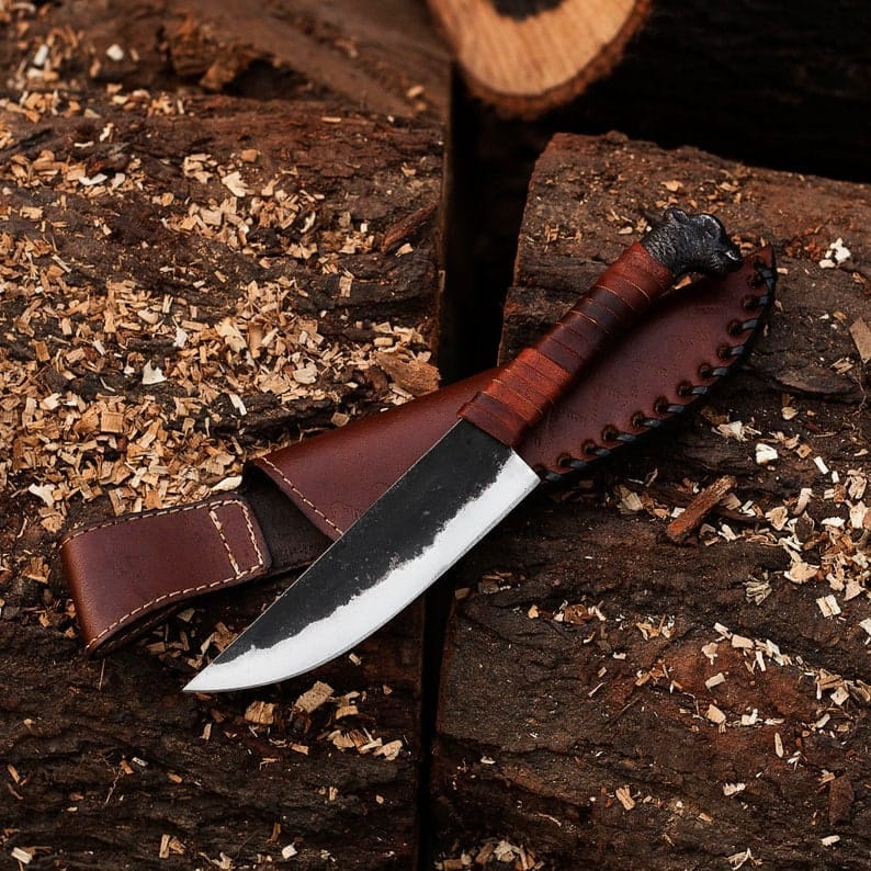 May include: A handcrafted knife with a dark blade and a light-colored edge, featuring a wrapped leather handle and a decorative animal head at the end. The knife rests in a brown leather sheath.