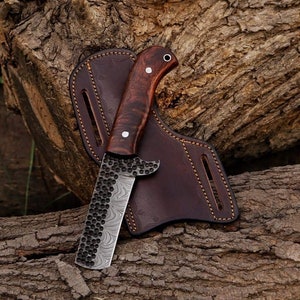 May include: A handcrafted knife with a dark wood handle and a patterned blade, resting in a brown leather sheath. The blade has a unique textured design. The knife is a tool for outdoor activities.