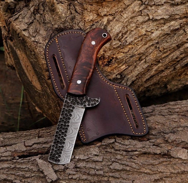 May include: A handcrafted knife with a dark wood handle and a patterned blade, resting in a brown leather sheath. The blade has a unique textured design. The knife is a tool for outdoor activities.