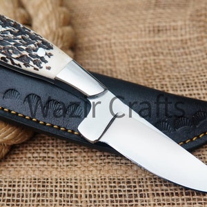 May include: A handcrafted hunting knife with a polished steel blade and a handle made of bone and dark brown material. The knife rests in a black leather sheath with the text "Wazir Crafts". The sheath is stitched with yellow thread.