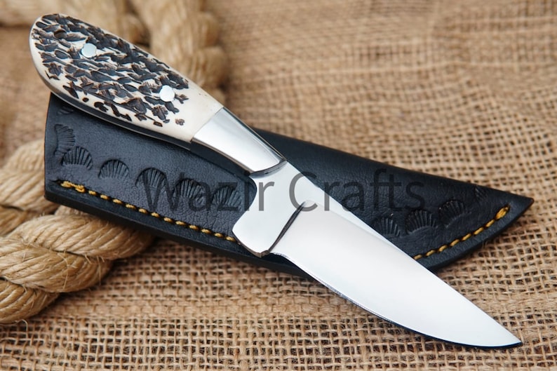 May include: A handcrafted hunting knife with a polished steel blade and a handle made of bone and dark brown material. The knife rests in a black leather sheath with the text "Wazir Crafts". The sheath is stitched with yellow thread.
