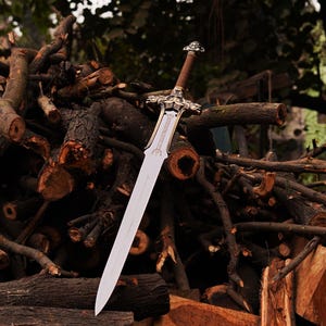May include: A double-edged sword with a silver-toned metal blade and a detailed crossguard featuring ornate carvings. The hilt is wrapped in brown material and topped with a metallic pommel. The sword is positioned vertically against a pile of wood.
