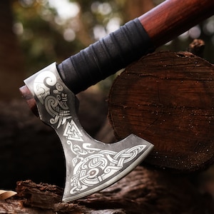 May include: A silver axe with a wooden handle and intricate etched designs. The axe is resting on a piece of wood.